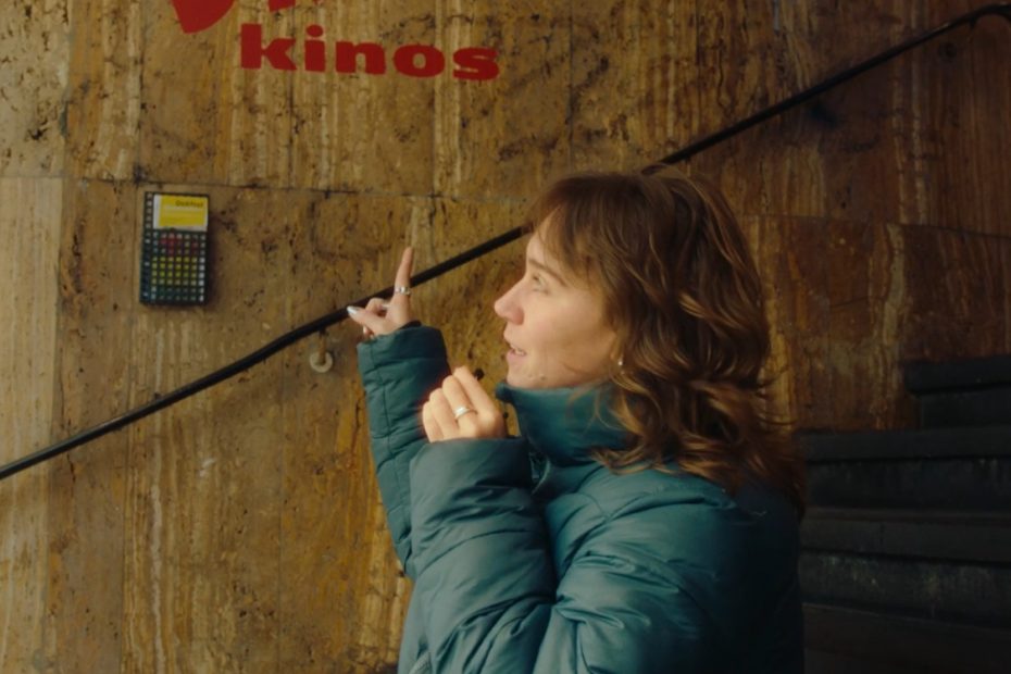 Person sitzt vor einer Wand und zeigt auf das Logo "Bali Kinos" hinter ihr